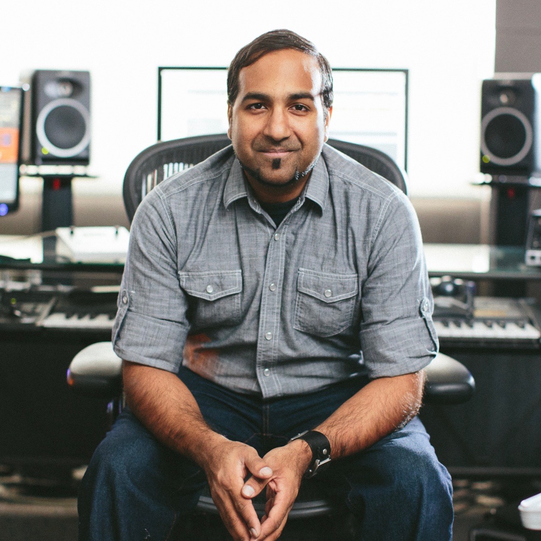 Portrait of Roy Oommen seated in his studio, monitors and keyboard behind him